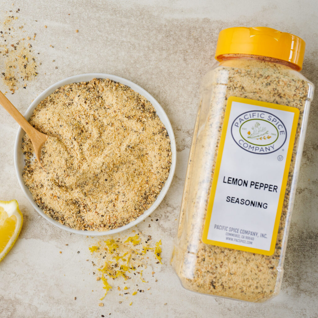 Pacific Spice Company lemon pepper seasoning in a white bowl with a wooden spoon, beside lemon wedges and the PSC1112 retail jar on a light surface